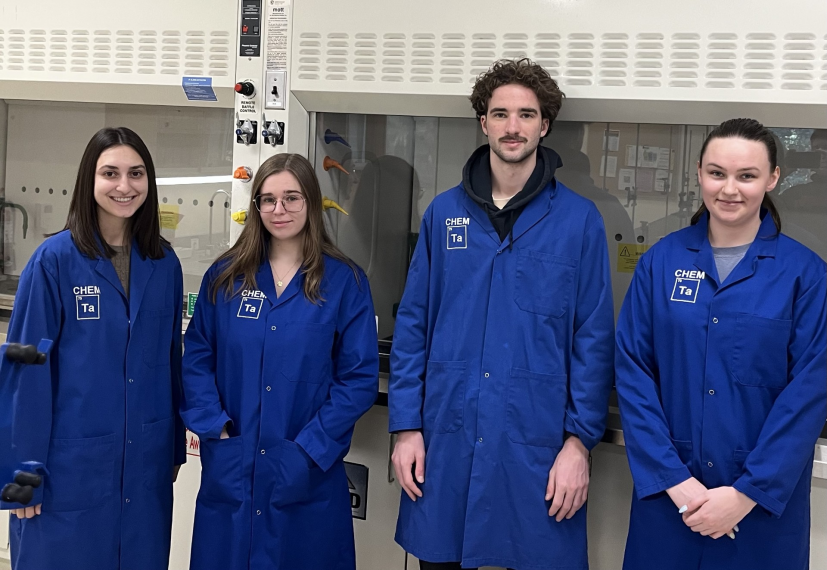 Four TAs stand in front of fumehoods in a laboratory. Each TA is wearing a blue lab coat.