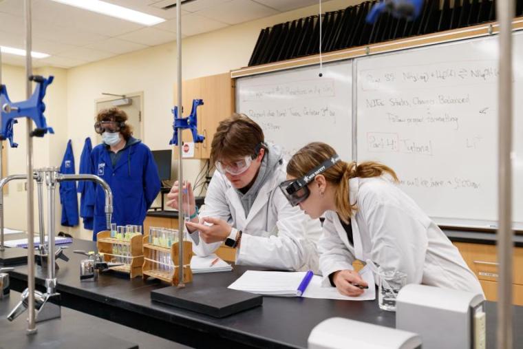 Students wearing lab coat and goggles observe a test tube with a clear colour liquid inside.