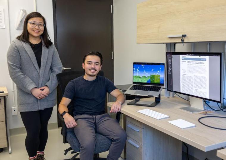 Prof. Leanne Chen stands next to Brendan Paget sitting in an office chair next to two computers. One screen displays Paget's artwork featured on a recently published journal.