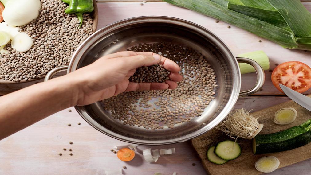 A photograph of a hand putting lentils in a pot, zucchini, a tomato slice and green pepper are on a cutting board nearby.