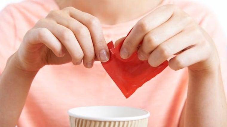 A woman in a pink shirt opening an artificial sweetener packet into her coffee.