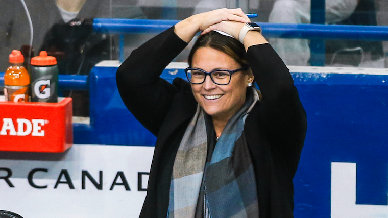 Rachel Flanagan, with her hands on top of her head, on the ice of a hockey rink, wearing a black coat and a blue tartan scarf.