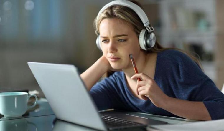 A woman at a desk, with a pencil, coffee cup and headphones, looking at a laptop screen.