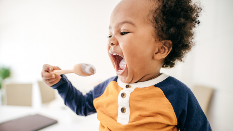 A toddler eating yogurt from a wooden spoon.