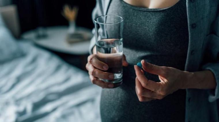 A photo of a pregnant woman, with a glass of water, taking a vitamin.