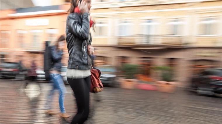 A photograph of a woman walking and talking on a cellphone on a sidewalk in a city.