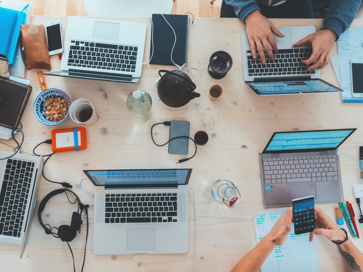 Bird's-eye view of a table covered in technology (laptops, cellphones, chargers, headphones) with folks working around the table.