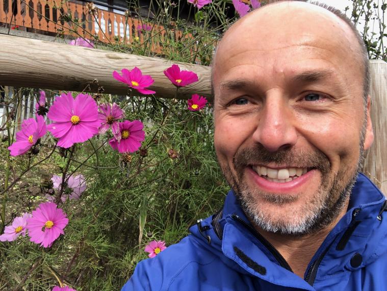 A photo of Dr. Steve Newmaster. He is wearing a blue jacket and standing beside pink flowers.