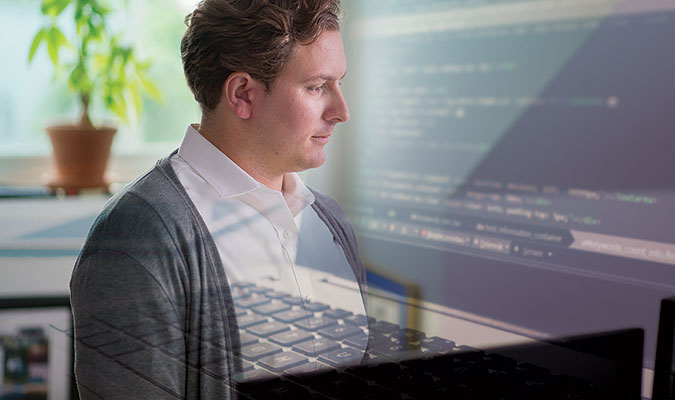 A young man with blonde hair wearing a white collared shirt and grey cardigan in an office space, with a window and potted plant out-of-focus in the background and an overlayed image of a computer screen and keyboard.