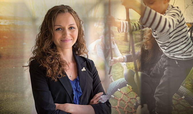 A young woman with shoulder-length, curly auburn hair with her arms folded, wearing a blue shirt and black blazer, smiling at the camera. Her image is overlayed by one of children playing in a tree-lined playground.