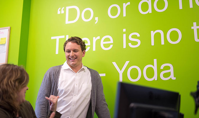 A man shows him smiling and standing in front of a vibrant green wall, mid-conversation with a female colleague who is sitting in front of a desktop computer.