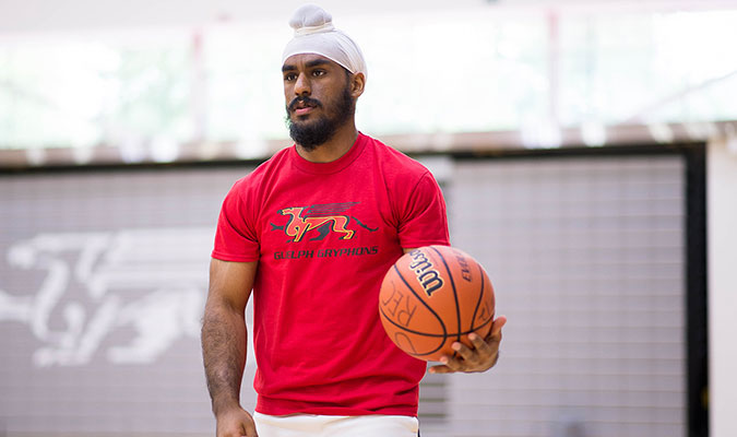 A young male student shows him in a red sports shirt with U of G's Guelph Gryphons logo emblazoned across the front, while he holds an orange basketball and looks to be walking across the basketball court floor.