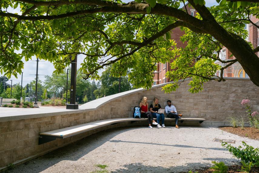 Three students in Lang Plaza sitting on a bench looking at a computer.