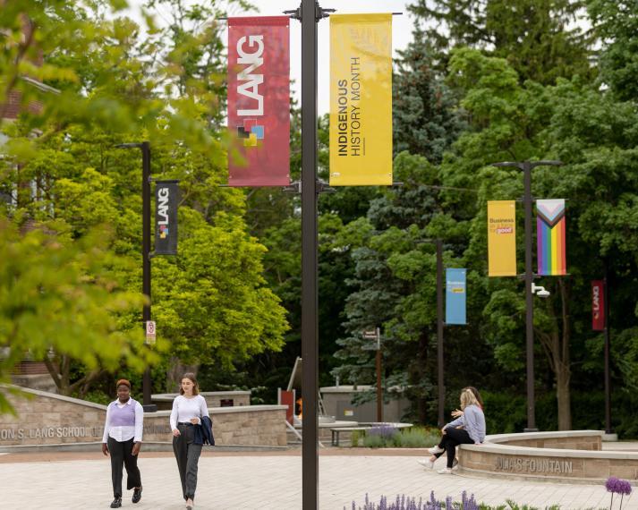 Students walking in Lang plaza with the Indigenous History Month flag and Pride flag in the background
