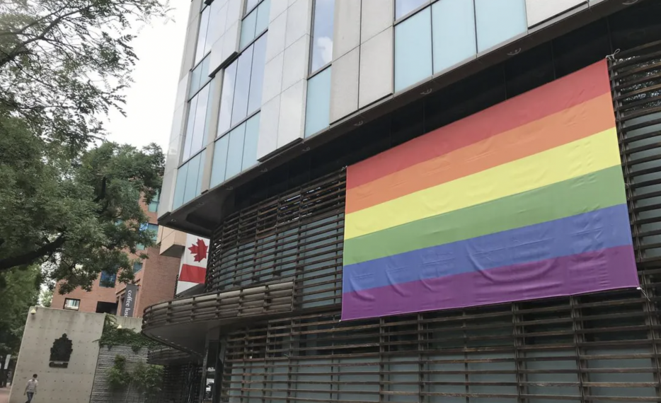 Pride flag hanging on a building with a Canadian flag in the background, less prominent.
