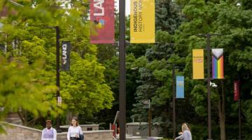 Students walking in Lang plaza with the Indigenous History Month flag and Pride flag in the background