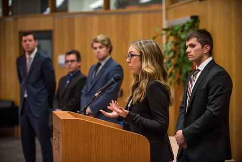 Female student speaks at podium with male counterparts looking on