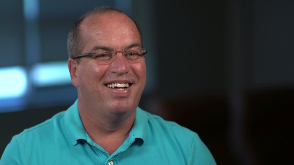 Chris Gillard smiling in a dark room; wearing a turquoise polo