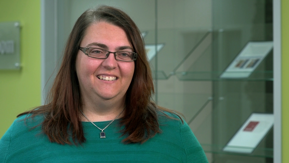 Prof. Katie Wood wearing green shirt standing in front of a glass book shelf