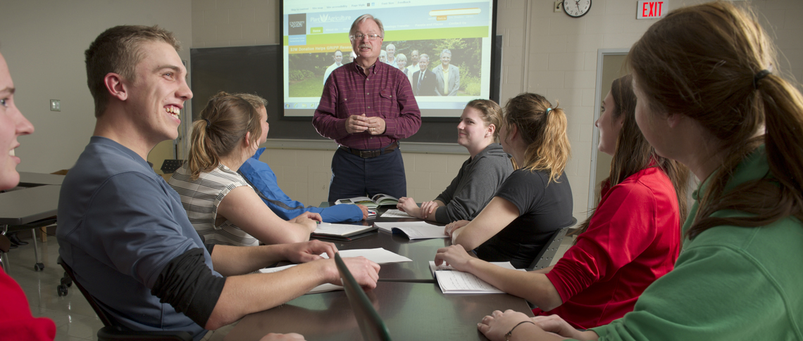 Group of students smiling while listening to an instructor at the front of a room presenting