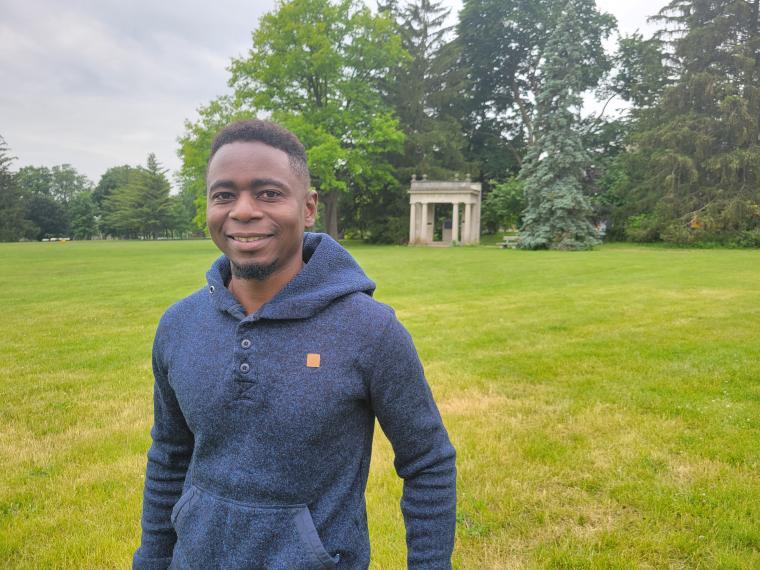 a photo of Taku wearing a blue sweater, standing on Johnson Green, with the Poritco behind him