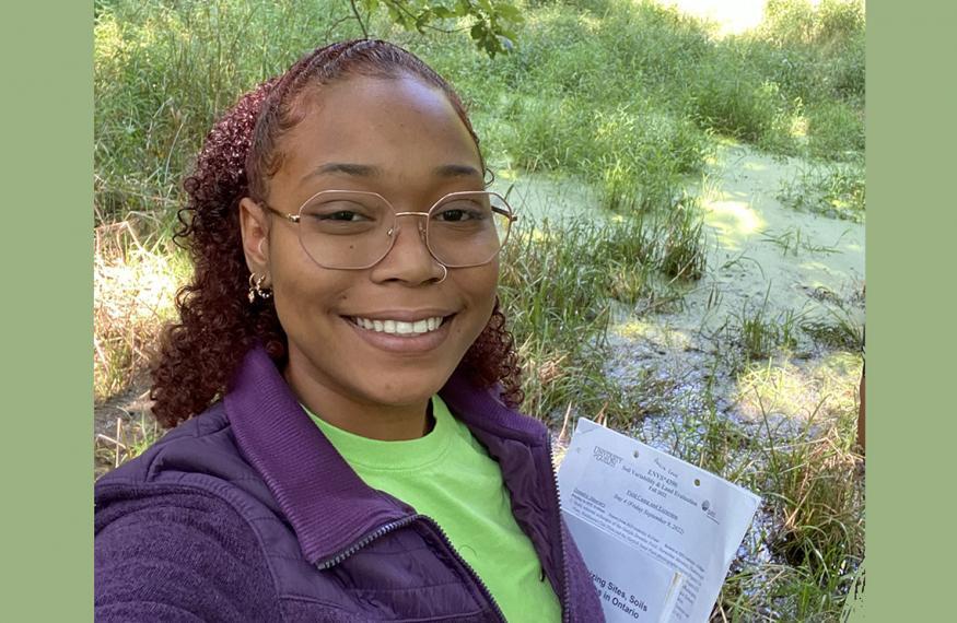 A selfie of Annia, in a green shirt, purple sweater and glasses, holding school work with sand and grass in the background.