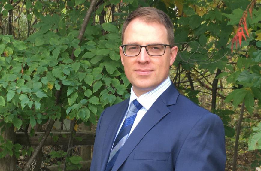 Brett Shepherd in a suit stands in front of green trees