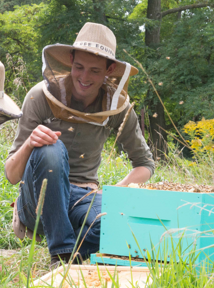 Head shot of Dan Borges, wearing a beekeeping hat, kneeling next to an open beehive brood box.
