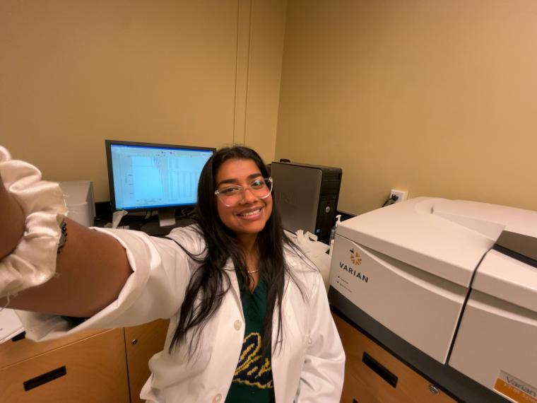 Vedika Nair sits in a chair with a lab coat on smiling.