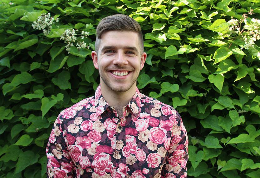 Jordan in pink floral dress shirt smiles in front of green foliage background