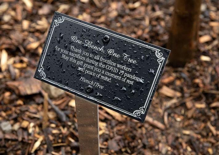 Black plaque with silver text that reads One Bench One Tree. Thank you to all frontline workers for your sacrifices during the COVID 19 pandemic. May this gift grant you a moment of rest and peace of mind. 1Bench1Tree.