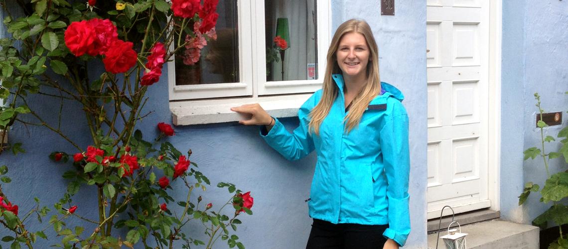 Emma Drake stands at window beside a rose bush.