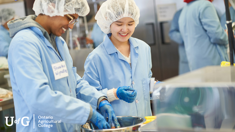 Two Food Science students stand in a lab with blue coats and hairnets on while smiling.