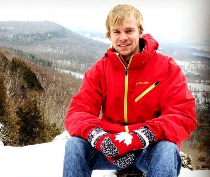 Jeremy sits at the top of a mountain wearing a red coat and mittens.