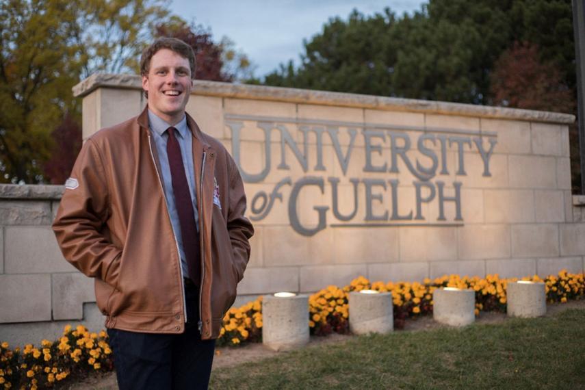 John stands with hands in pockets of brown leather jacket in front of University of Guelph sign