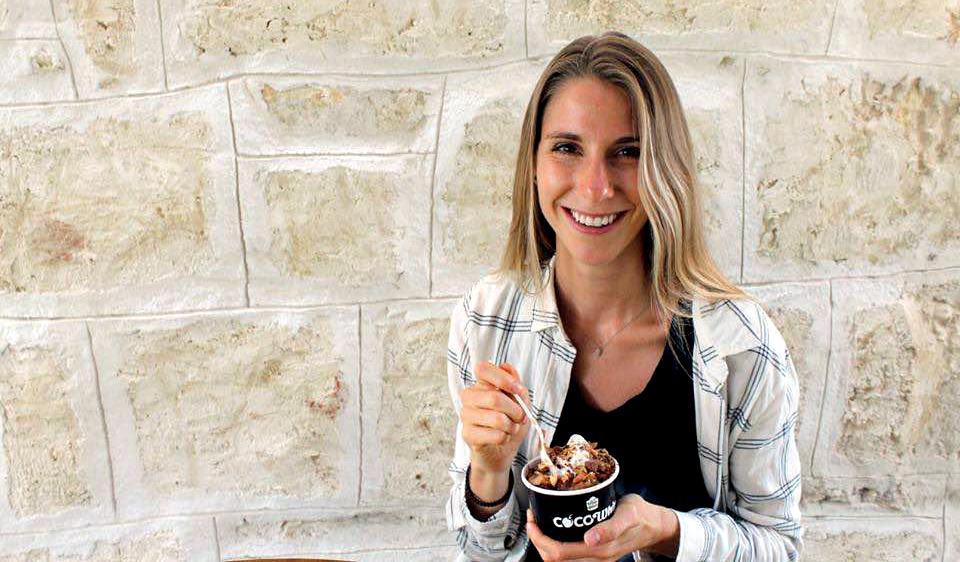 Kristi stands in front of a stone wall with a cup of food and a fork in her hand.