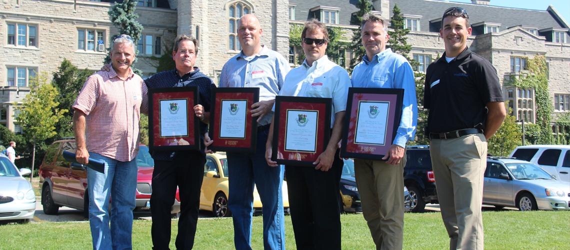 Six men stand in front of Johnston Hall outside, four award winners hold framed certificates