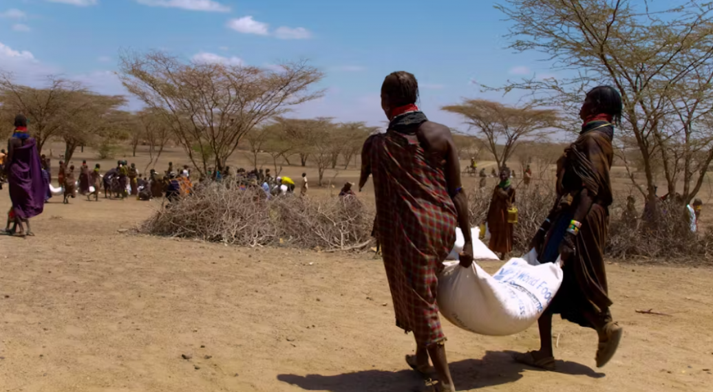 Two women carrying a sack of food between them while walking towards a larger group of women doing the same thing in a dry, arid area