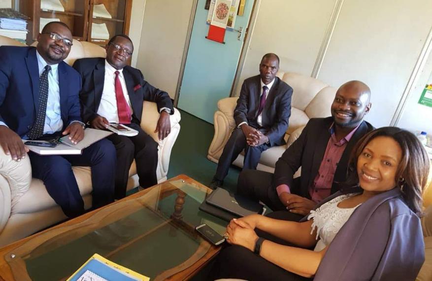 Group of sitting government officials smiling, Tendai sitting with them on the right.