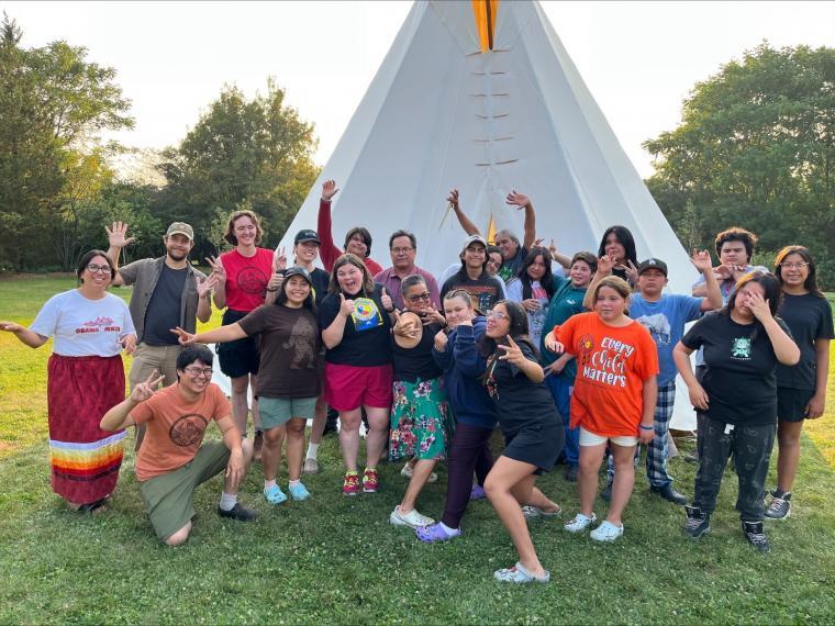 A group of people pose playfully in front of a tipi outdoors.