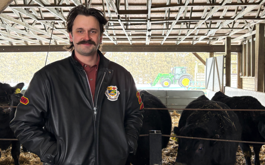 wyatt standing in a barn with cows and a tractor behind him. he's wearing a black leather jacket with the oac crest on it
