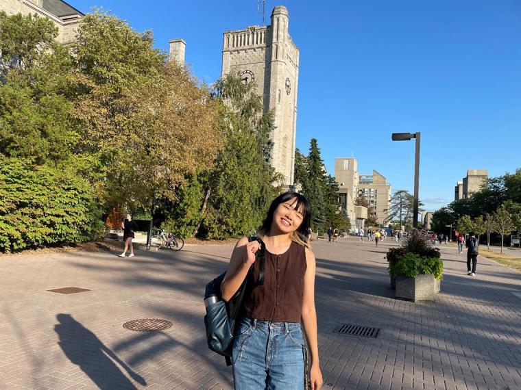 A woman wearing jeans, a brown shirt and a bakcback posing in front of a historic brick buidling on a bright summer day.