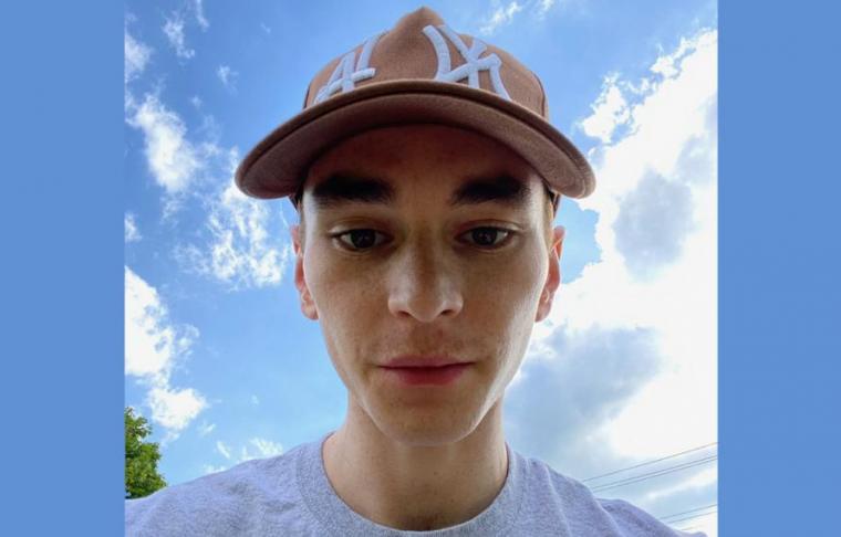 A selfie of Benjamin, wearing a grey t-shirt and brown baseball hat, with blue sky in the background.