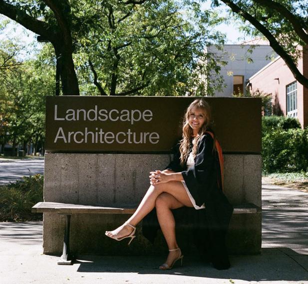 A woman with long hair dressed in heels and graduation gown sitting on a bench in front of a landscape architecture sign.