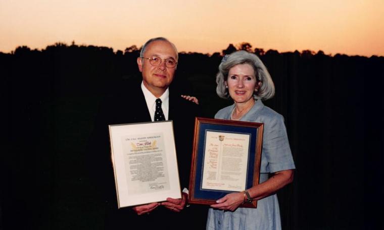 Jane and Tom Funk stand behind a sunset smiling while holding up their Distinguished Teaching Awards (from the OAC)).