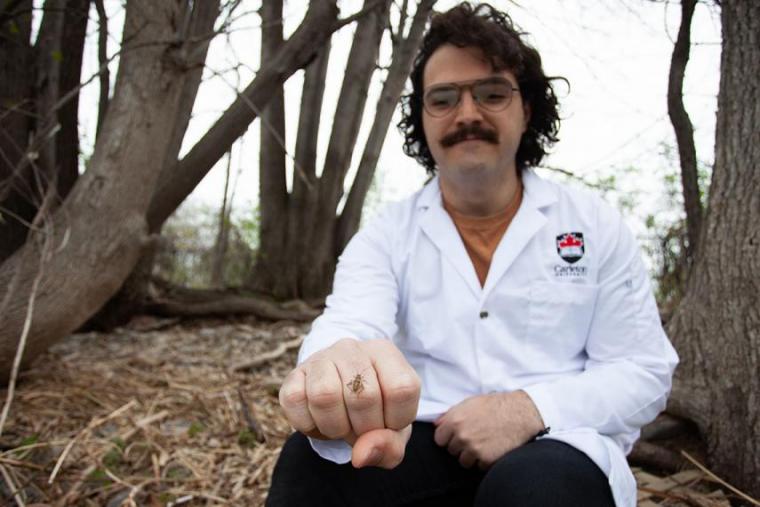 Matt with white lab coat on sits outside in woods with a cricket on his hand