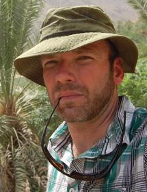 Headshot of Andrew Burton Anderson wearing hat and holding sunglasses in mouth.