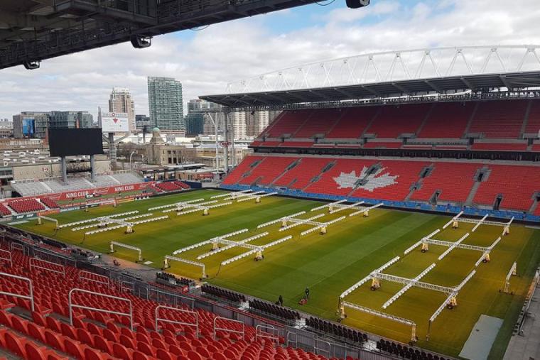 Grass field surrounded by red stadium seats; large light fixtures over the grass