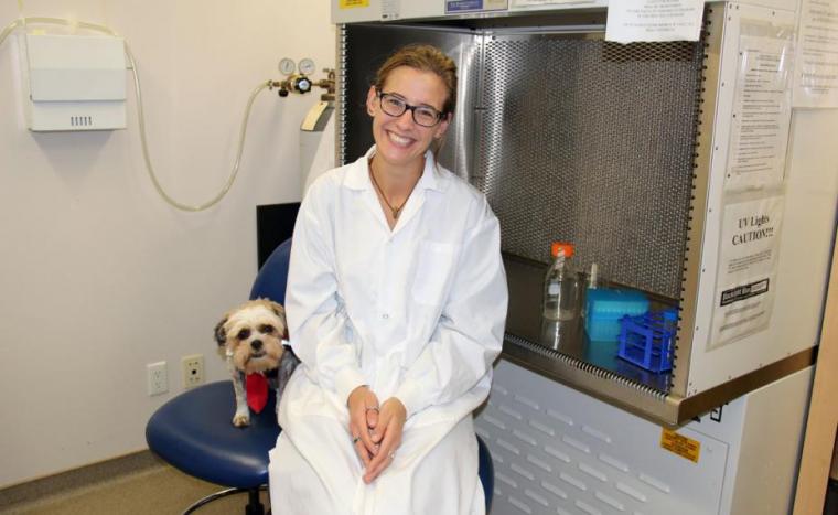 Jennifer in lab coat sits in lab with small dog sitting beside her