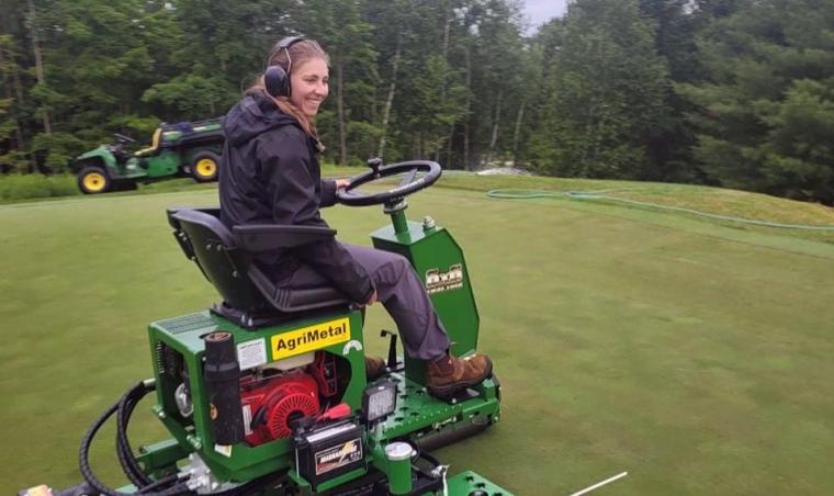 Katie on a small lawn tractor smiling as she drives on a golf course